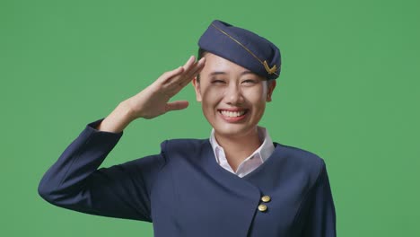아시아 여성 승무원이 녹색 스크린 배경 스튜디오에 서서 경의를 표하고 인사를 하고 있습니다.