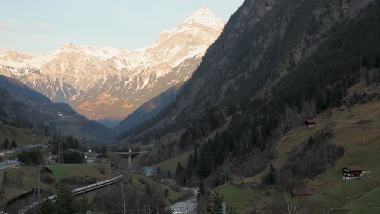 valle alpino con montañas cubiertas de nieve, un tren curvado a lo largo de las vías, carretera y casas esparcidas al anochecer