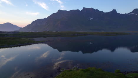 Low-aerial-flyover-of-a-van-parked-near-a-calm-lake-with-a-beautiful-reflection-of-the-mountain-and-sky-in-the-early-evening-as-the-sun-is-setting