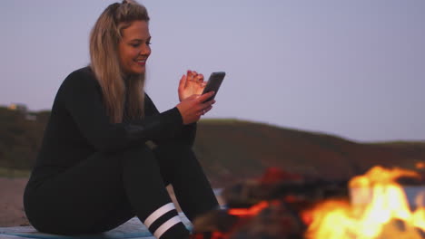 woman sitting on surfboard by camp fire on beach using mobile phone as sun sets behind her