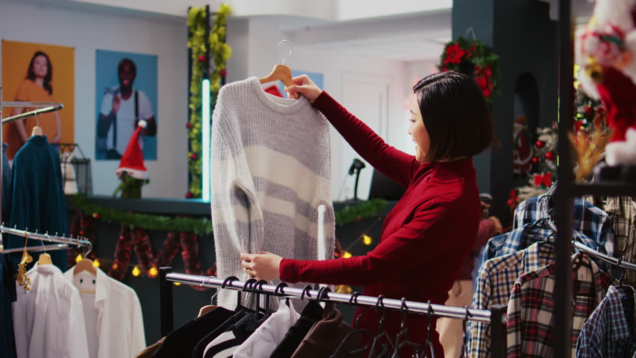 명랑한 아시아 고객은 크리스마스 화려한 옷가게에서 명절 연휴 기간 동안 크리스마스 판촉 세일을 이용하여 통관으로 편안한 옷을 사려고 합니다.