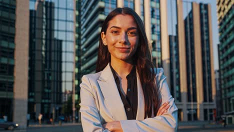 joven mujer de negocios segura de pie con los brazos cruzados cerca de rascacielos, encarnando el éxito profesional durante la hora dorada en el distrito financiero urbano, irradiando ambición de carrera