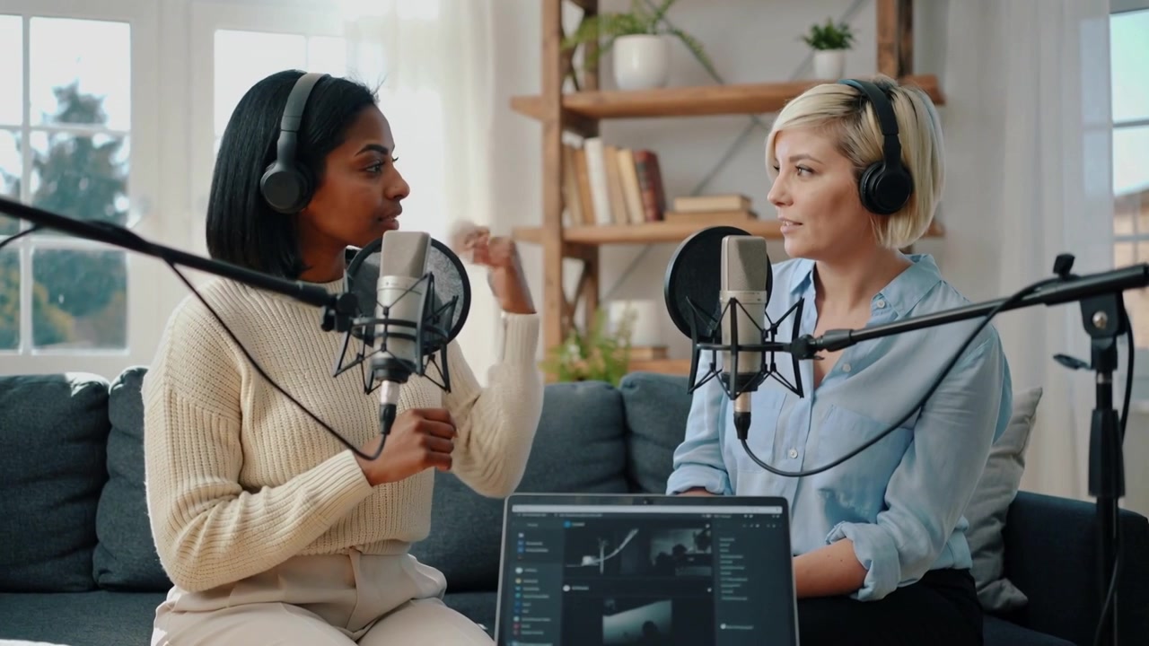 Leaning, left host speaking and gesturing to cohost listening in home studio, recording with mics