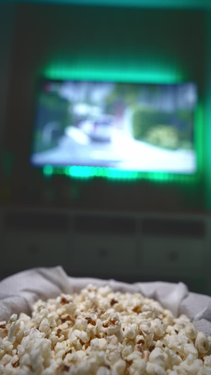 Primo piano di una mano che afferra popcorn fatti in casa da una ciotola con un film in riproduzione sullo sfondo, a illustrare una accogliente serata film a casa, spuntini e intrattenimento casuale.