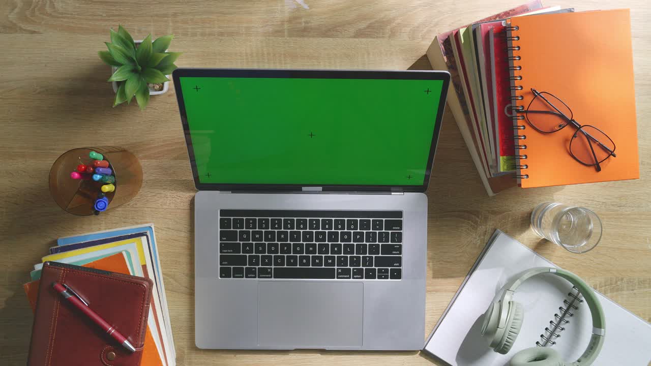 vista de arriba hacia abajo de una computadora portátil con pantalla verde simulada pantalla chromakey en un escritorio de oficina de madera al lado de la libreta con bolígrafos, gafas y un vaso de agua.