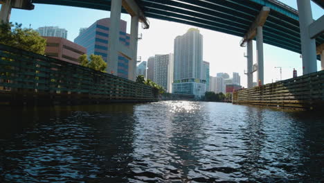 view-from-a-small-watercraft-as-it-passes-beneath-a-bridge-in-the-waterways-of-Miami-Florida-to-reveal-tall-buildings-and-a-bright-sun