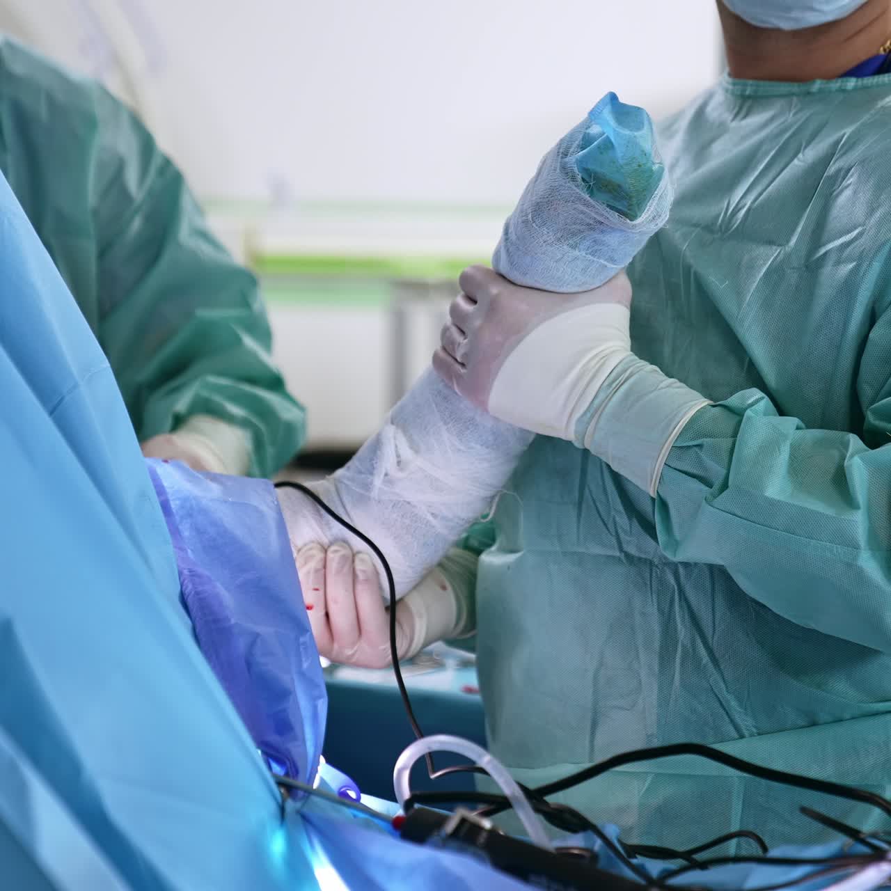 Assisting doctor holding a patient's hand covered with bandaging during operation. Main doctor performing operation at backdrop