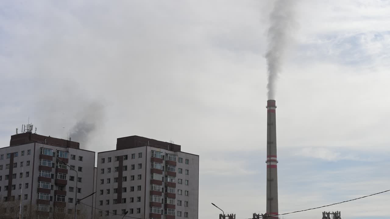 A tall industrial chimney emits smoke next to residential buildings in Ulaanbaatar, Mongolia. The power plant infrastructure looms over the city's living quarters and skyline
