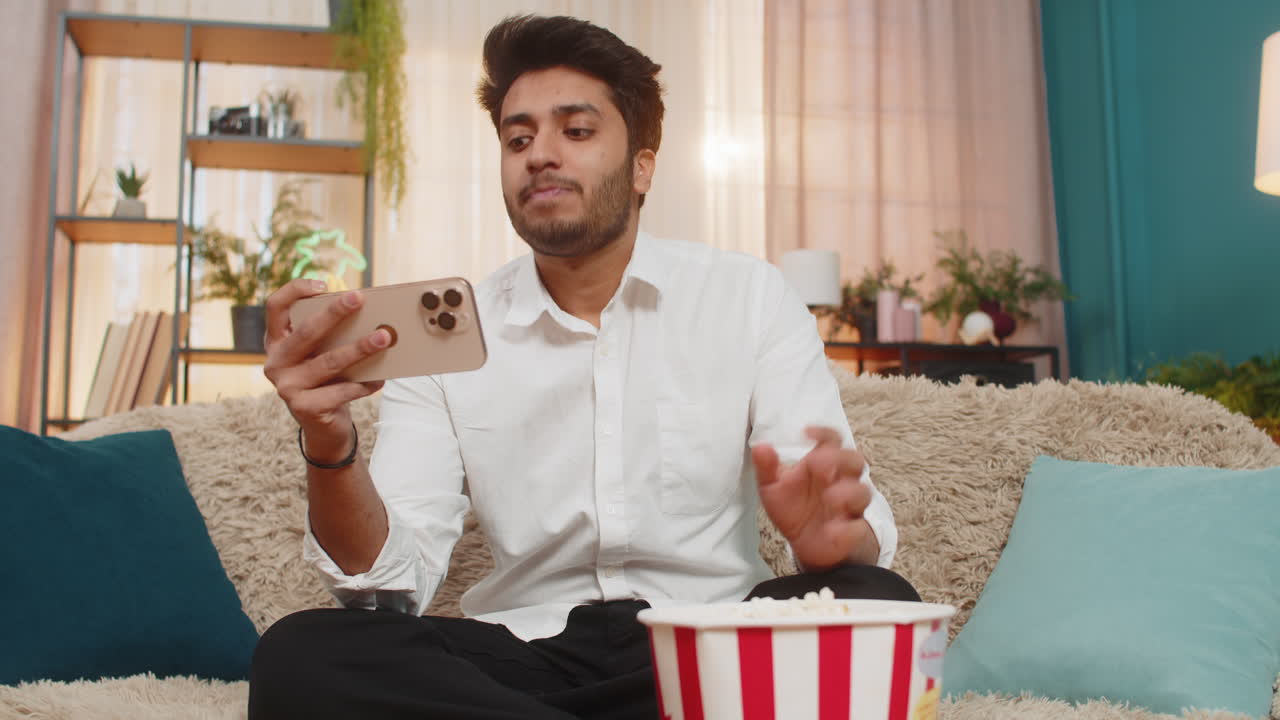 Indian man sitting on sofa eating popcorn watching video on smartphone focused enjoying relaxation