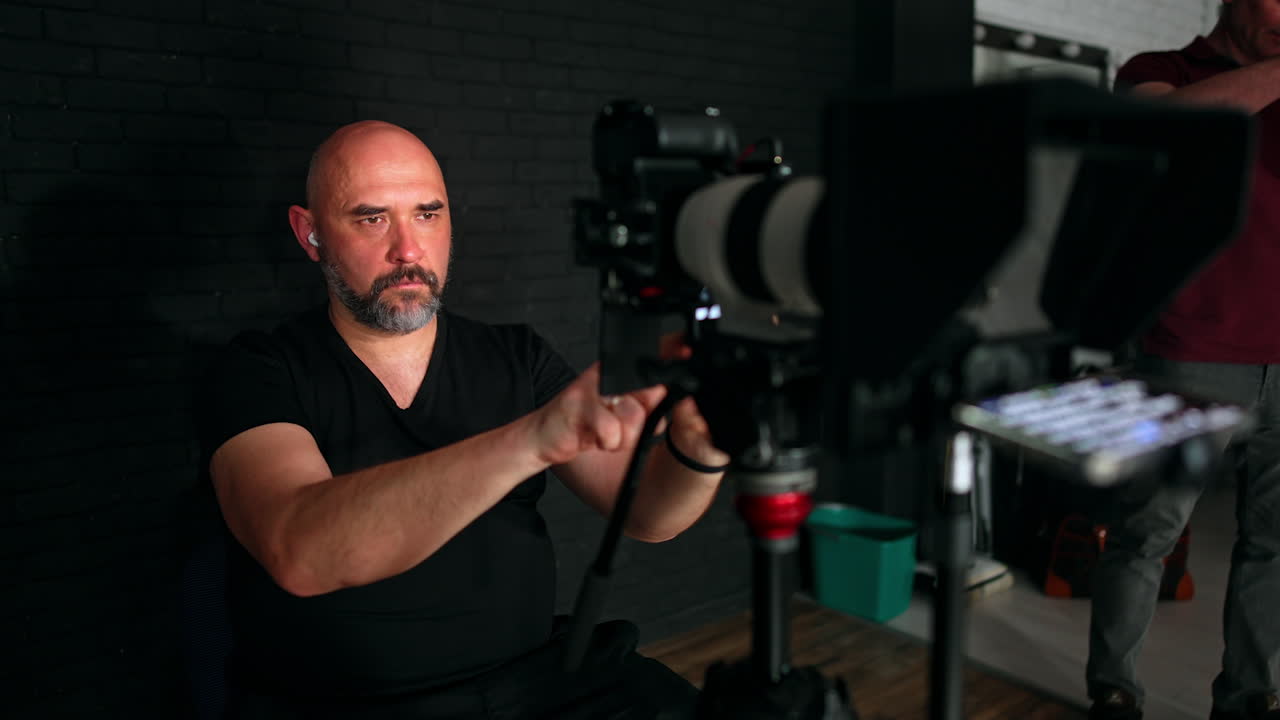 Focused bearded bald photographer sits on the chair. Cameraman looks at display. Work in studio.