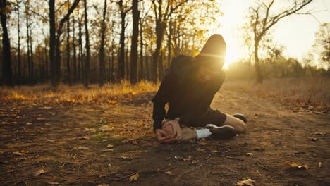 A-man-in-a-black-sports-uniform-with-a-red-cap-makes-a-face-and-holds-his-knee-while-jogging-in-the-forest-he-lies-on-the-ground.-Morning-jogging-safety-rules