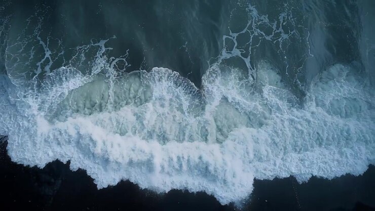 Aufschäumende Meereswelle bricht auf dunkle Küste, erzeugt spitzenartiges Weißwasser und Schaum.