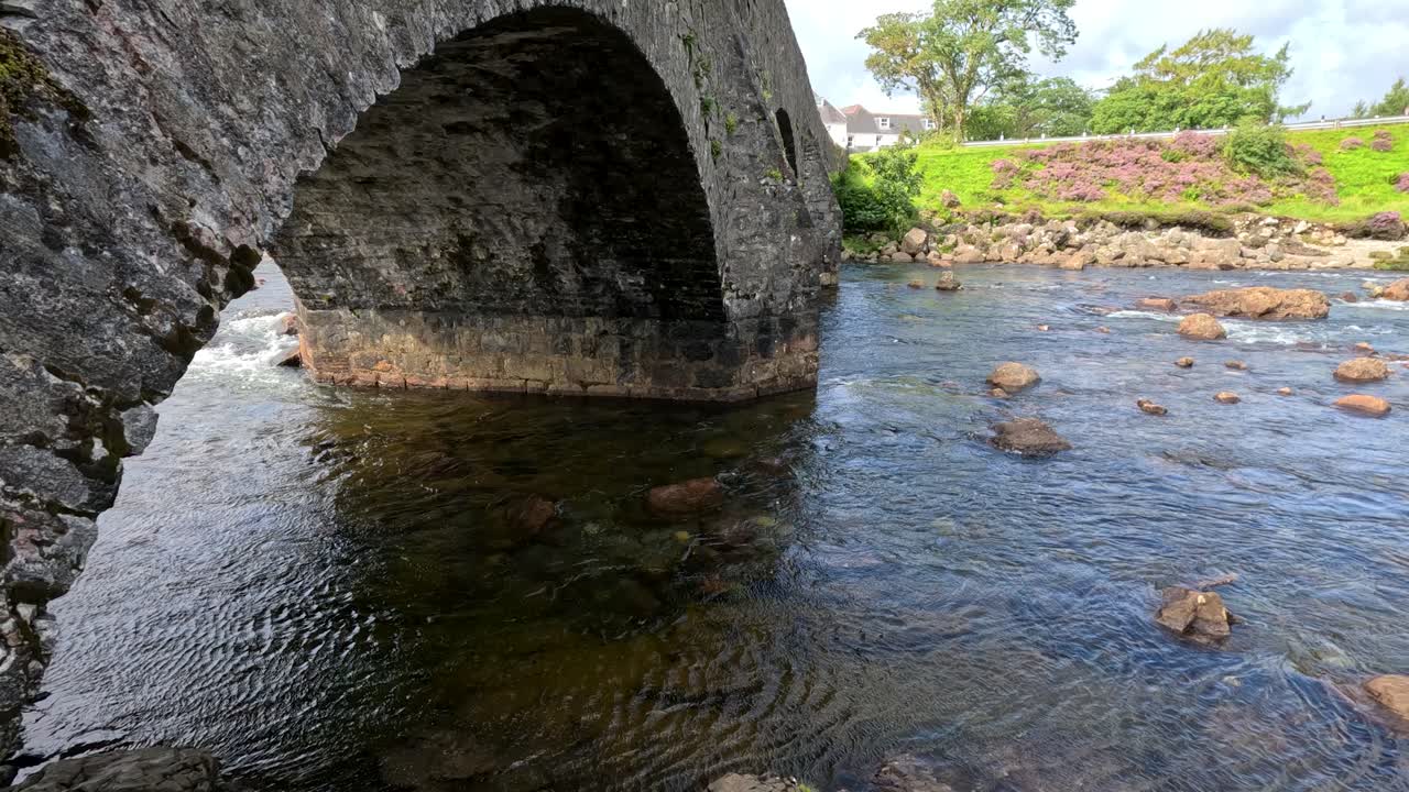 Camera slowly pans beneath a historic stone arch bridge, revealing clear river water, scattered rocks, and lush green landscape in natural daylight