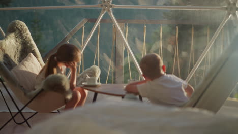 children observe nature outside glamping tent. calm siblings sit at table at beginning of new day on summer vacation. bored kids in hotel dome