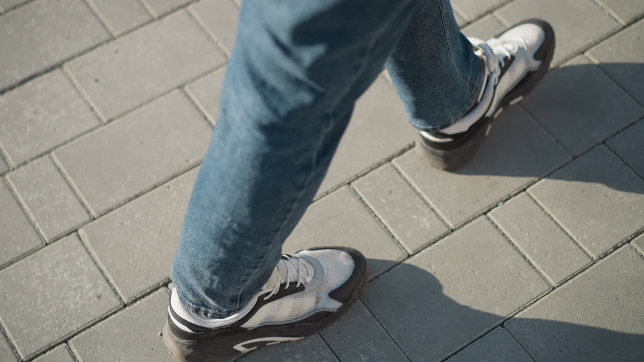 Top-down close up of person in jeans and canvas sneakers walking on interlocked pavement tiles during sunny day with long shadow cast on clean ground