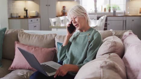 una mujer caucásica anciana feliz sentada en el sofá de la sala de estar, usando una computadora portátil y hablando en su teléfono inteligente.