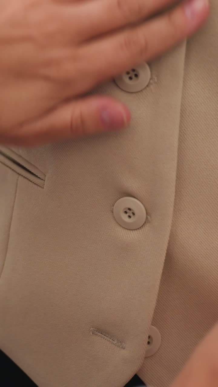 Rematador caucásico inspeccionando los detalles de los botones, primeros planos meticulosos de los dedos que alinean el ojal y aseguran las puntadas, textura de tela neutra, ambiente de control de calidad en un taller moderno.