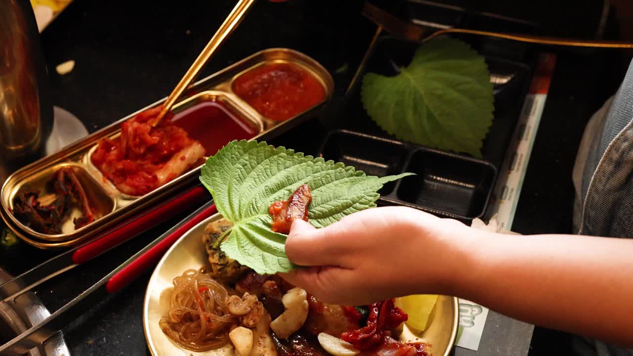 Hand places grilled pork and kimchi on perilla leaf, overhead view, warm restaurant lighting