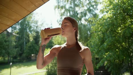 A-blonde-girl-in-light-brown-in-a-T-shirt-opens-a-special-thermos-and-drinks-water-against-the-backdrop-of-a-green-forest-in-the-gazebo