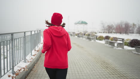 rücksicht auf eine frau, die in roter mütze und jacke entlang eines schneebedeckten weges in der nähe eines eisenrails joggt, ruhige winterpark-umgebung, bänke in der nähe, neblige atmosphäre mit fernen brücken und schneedeckten bäumen