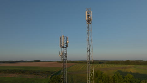 Two-radio-towers-in-the-middle-of-farmland-during-sunrise,-aerial-orbital