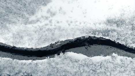 descending aerial top down of frozen river surrounded by white winter landscape with snow during bright day