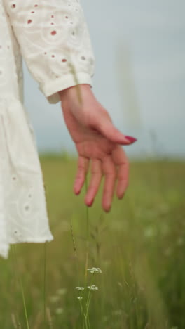 un primer plano de la parte inferior del cuerpo de una mujer mientras se pasea por un campo verde exuberante, con un vestido blanco que fluye. su mano toca suavemente la hierba alta