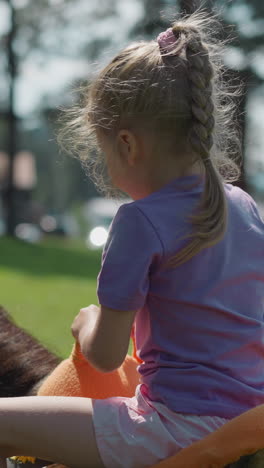 cuidadosa niña rubia con camiseta lila monta en pony con un instructor experto a lo largo del jardín verde de la ciudad en un día soleado vista trasera en cámara lenta