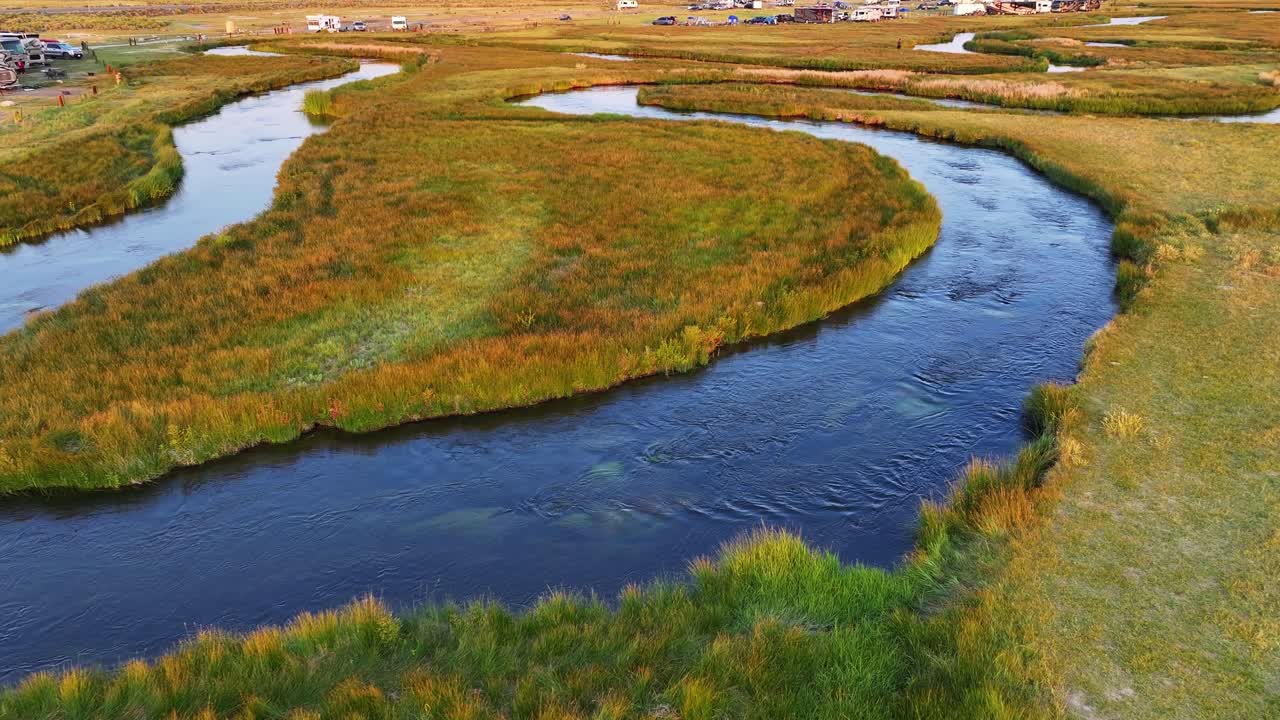 Matalalla lentävä drone lähestyy mutkittelevaa Owensjokea vihreiden niittyjen ja rantojen läpi lähellä Mammoth Lakesia Kaliforniassa lämpimässä iltavalossa. Tasainen lähestyminen korostaa S-mutkan muotoisia vesikanavia