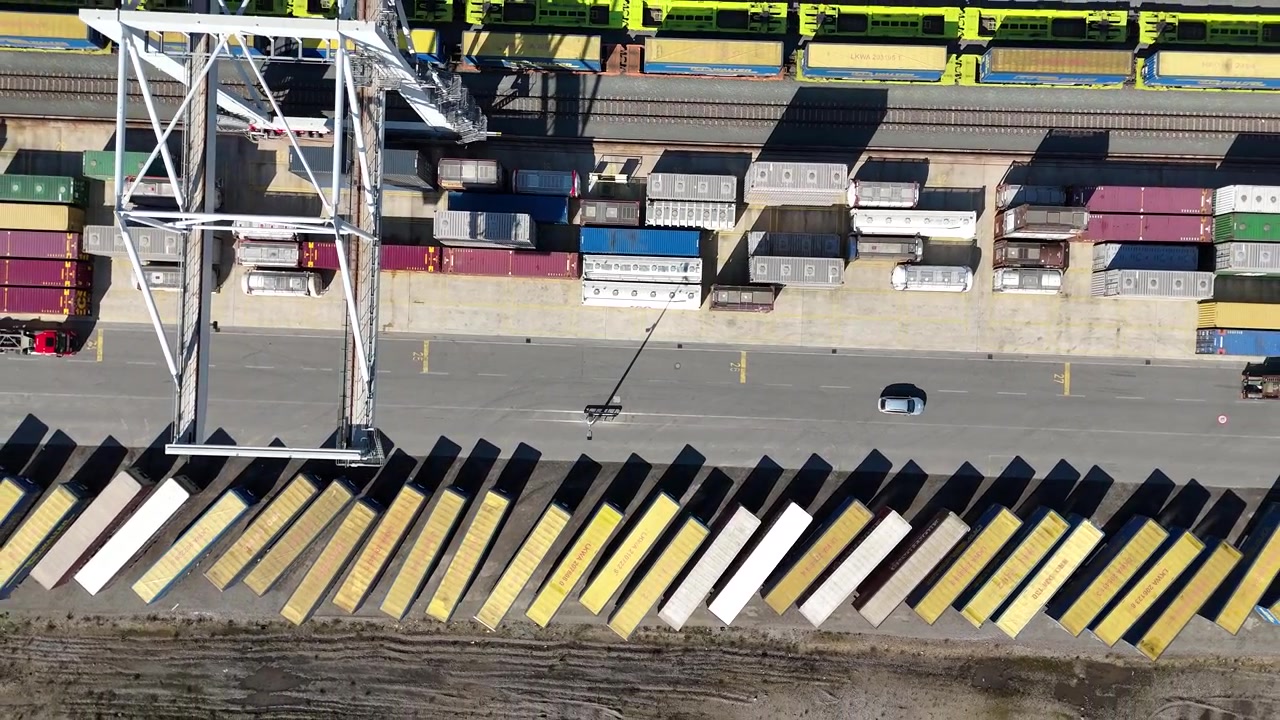 Drone lifts upward above stacked containers as a large crane slides across the terminal and a car passes below.