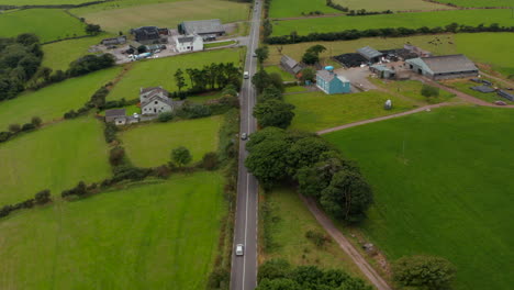 Blick-Aus-Der-Vogelperspektive-Auf-Autos,-Die-Auf-Einer-Geraden-Straße-Fahren,-Die-Um-Bauernhöfe-Herumführt.-Nach-Oben-Kippen-Geben-Den-Panoramablick-Auf-Die-Landschaft-Mit-Feldern-Und-Weiden-Frei.-Irland