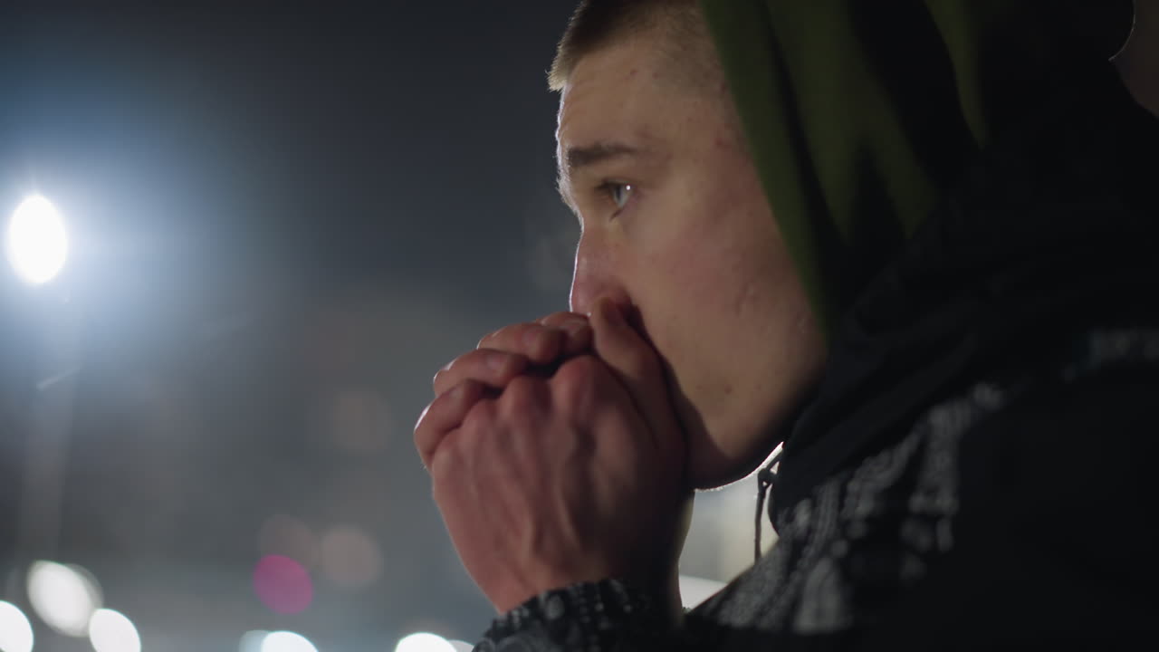 Cold man in hoodie exhales warm air onto hands for warmth as cold vapor rises, captured against blurred night background with distant light illuminating winter atmosphere