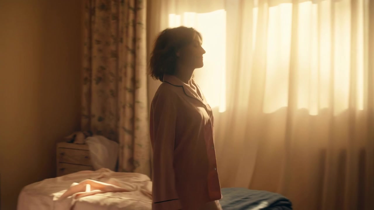 Camera da letto illuminata dal sole al mattino, donna seduta sul letto in abiti rosa, che guarda verso la finestra mentre filtra una calda luce dorata