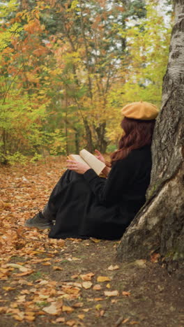 vista lateral de una dama hojeando un libro pensativamente mientras se apoya en un árbol, las hojas de otoño cubren el suelo mientras disfruta de la tranquila soledad en la naturaleza serena, absorta en páginas de conocimiento