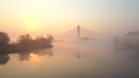 low flying over a wide river, towards a modern cable bridge during a foggy sunrise morning