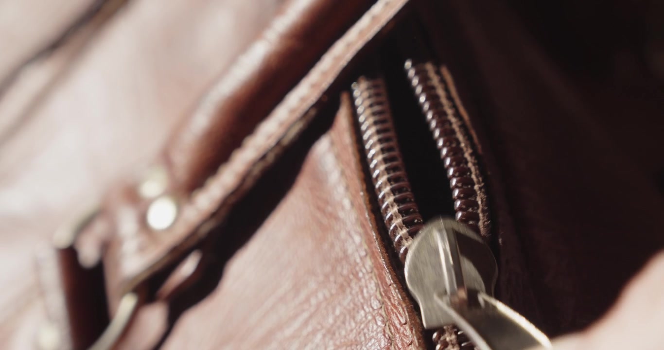 Un dettaglio macro mostra una vecchia borsa da viaggio in pelle usurata che viene aperta con la cerniera alla luce del sole naturale, creando uno sfondo nostalgico di viaggio.