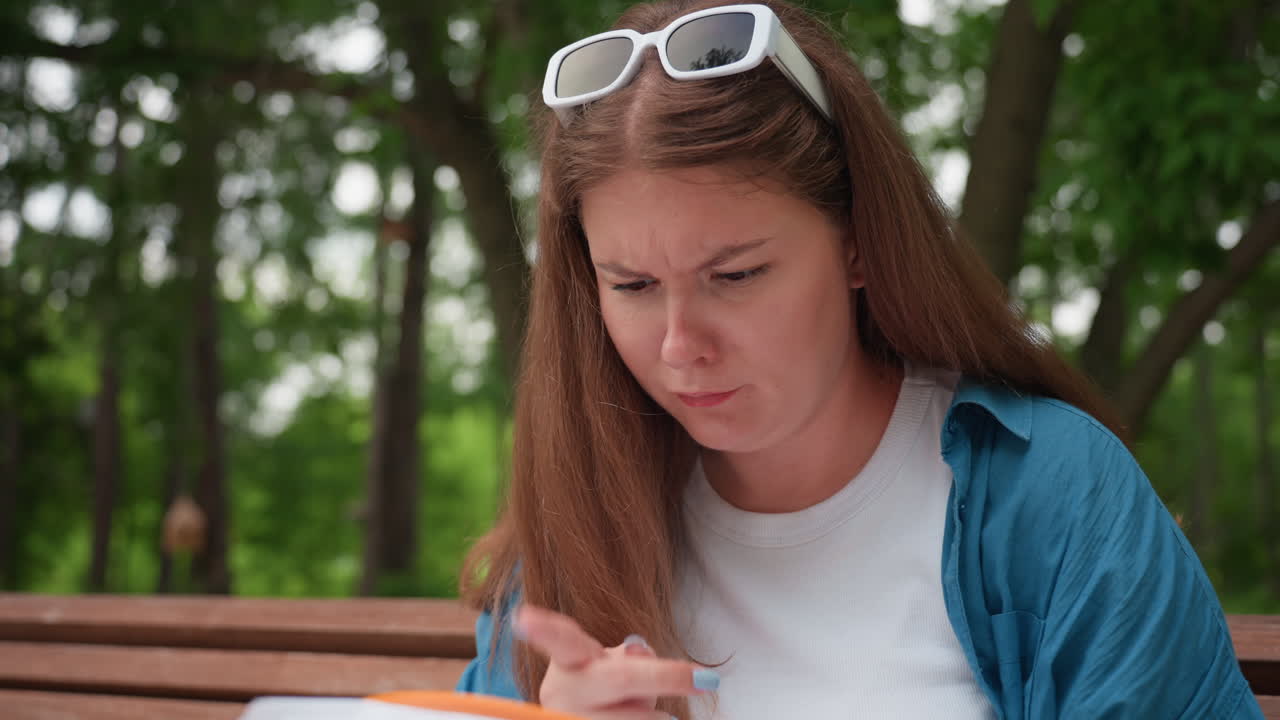 Una joven sentada en un banco del parque observa de cerca un bordado mientras señala el diseño con expresión de disgusto, rodeada de exuberantes árboles verdes y una cálida luz diurna.
