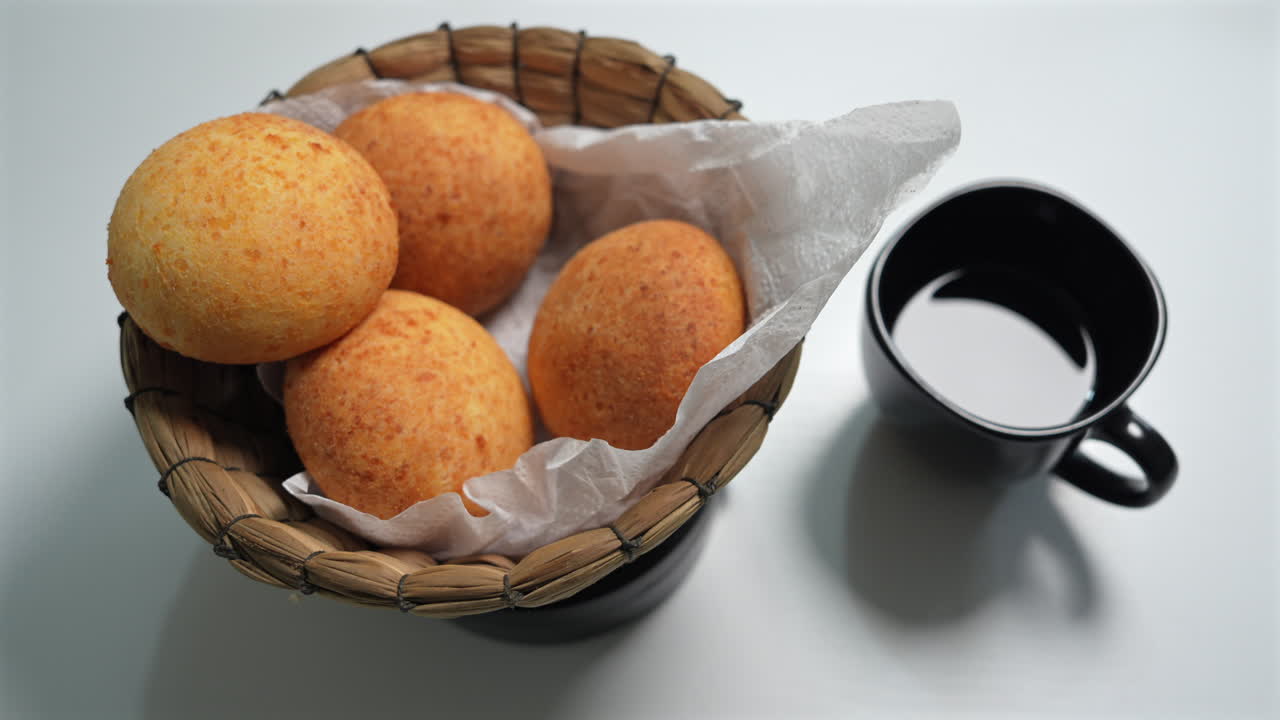 Rustic basket of golden almohabanas beside a black coffee cup on a white table. Bright lighting enhances the cozy, inviting breakfast scene in close-up shot