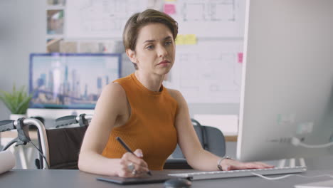 Disabled-Female-Architect-In-Wheelchair-In-Office-Working-On-Desktop-Computer-Using-Graphics-Tablet