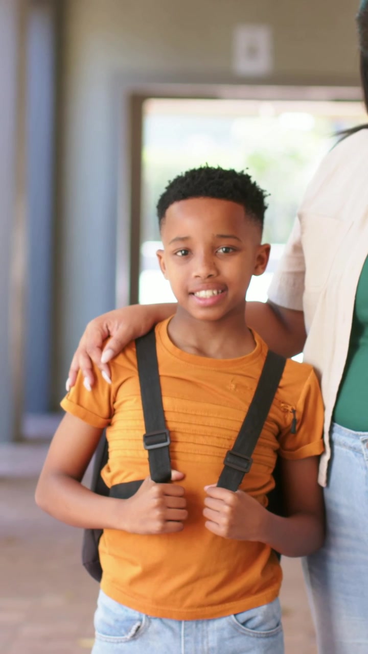 Posing boy in orange tee holding black backpack straps at building entry guardian arm on shoulder