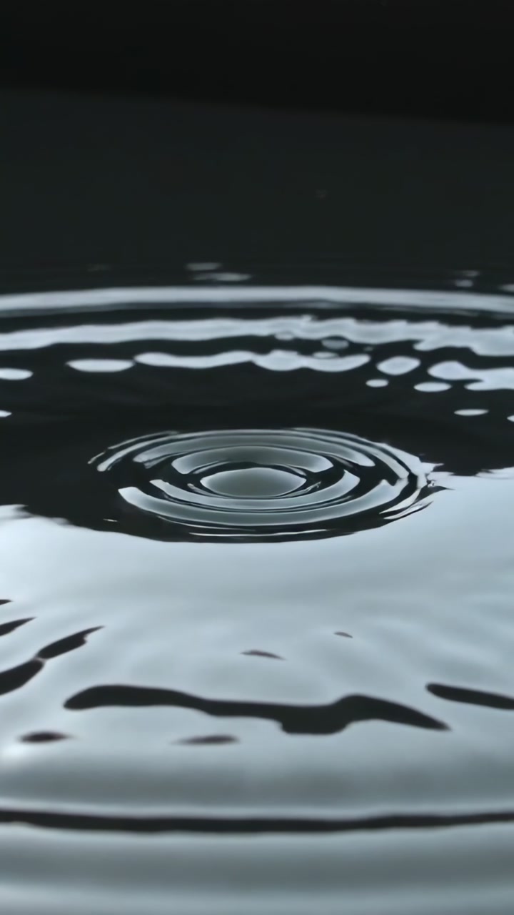 Vertical video: Droplet striking shiny pool, forming crown, rebound dome and ripples in studio