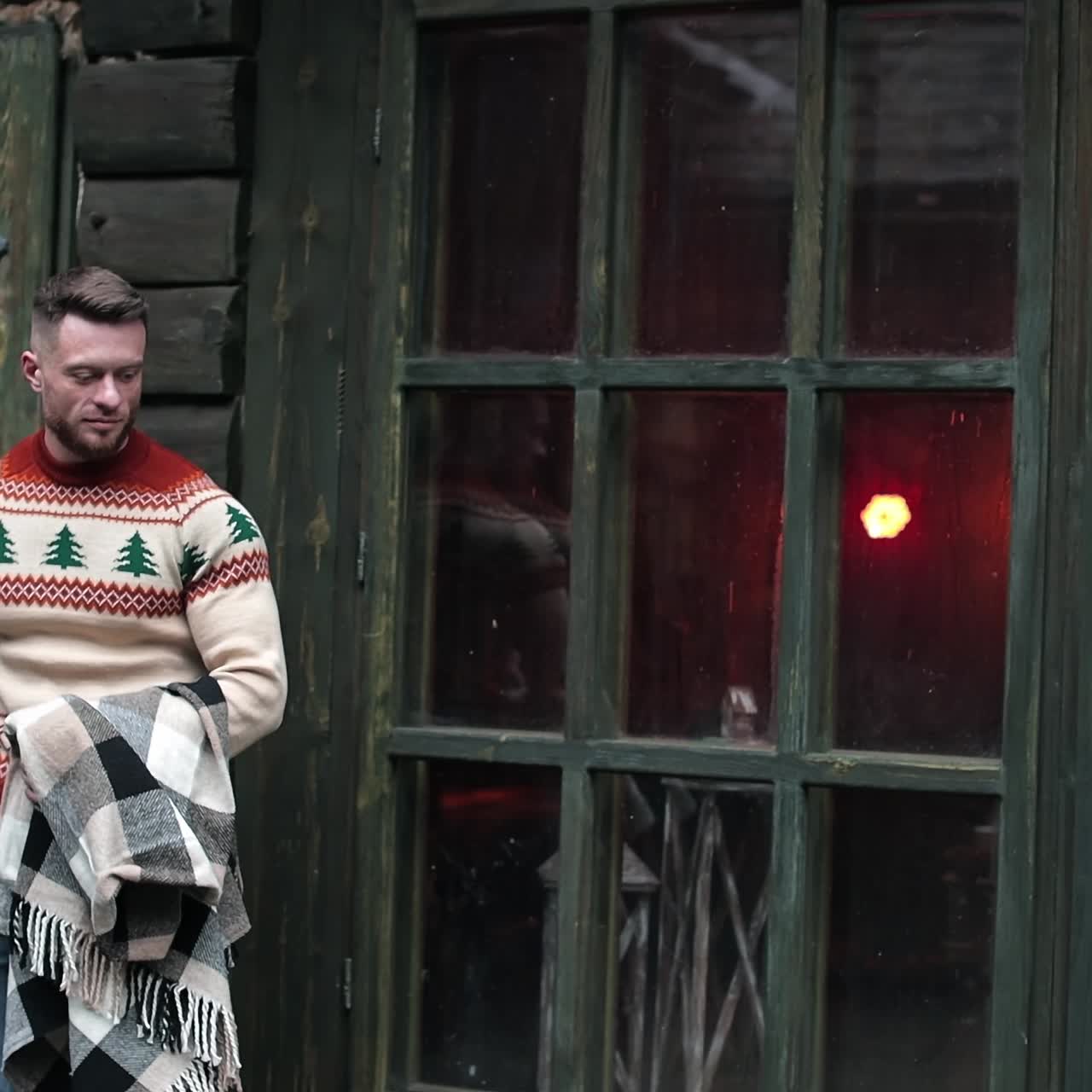 Handsome bearded man walks out of the old rural house. Fit man in sweater goes somewhere holding warm plaid. Attractive man at wooden house background