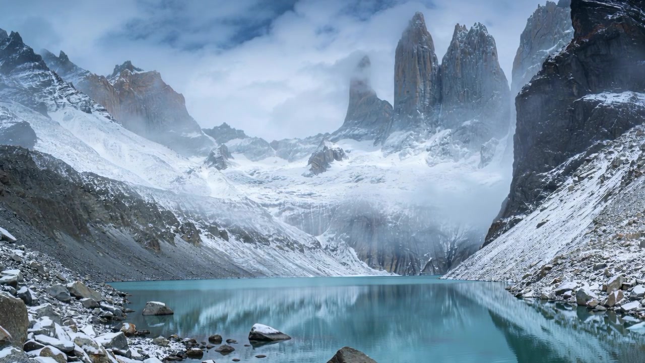 Nuvole basse e nebbia che si muovono, rivelando cime innevate su un lago turchese, il vento crea increspature