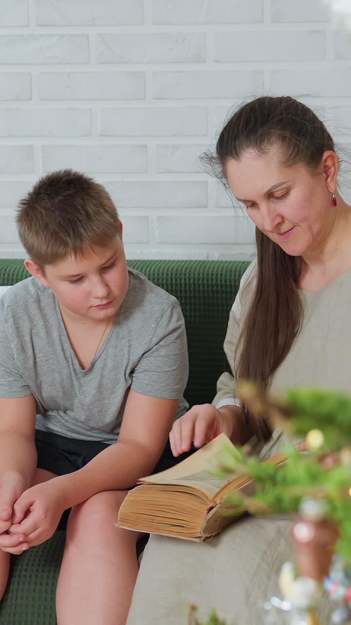 Madre e hijo blancos leyendo un libro en un sofá verde contra una pared de ladrillo blanco, madre guiando a su hijo a través de un álbum de fotos familiar antiguo, niño atento pasando las páginas, luz natural cálida, aprendizaje doméstico tranquilo