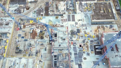 Aerial-Bird-Eye-View-Of-a-Construction-Site-Building-Cranes-Looking-Down-Industrial-Machinery-Area-around-Residential-Urban-Apartments