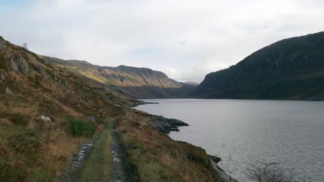 Looking-down-a-remote-hiking-trail,-sunlight-highlights-the-distant-mountain-slopes-of-a-dark-Scottish-glen-filled-with-a-large-sea-loch-and-surrounded-by-tall-cliffs