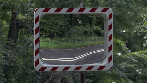 A-convex-mirror-positioned-at-a-road-junction-in-a-forest-reflects-the-road-and-passing-vehicles-as-a-safety-feature-at-a-road-junction,-Worcestershire,-England