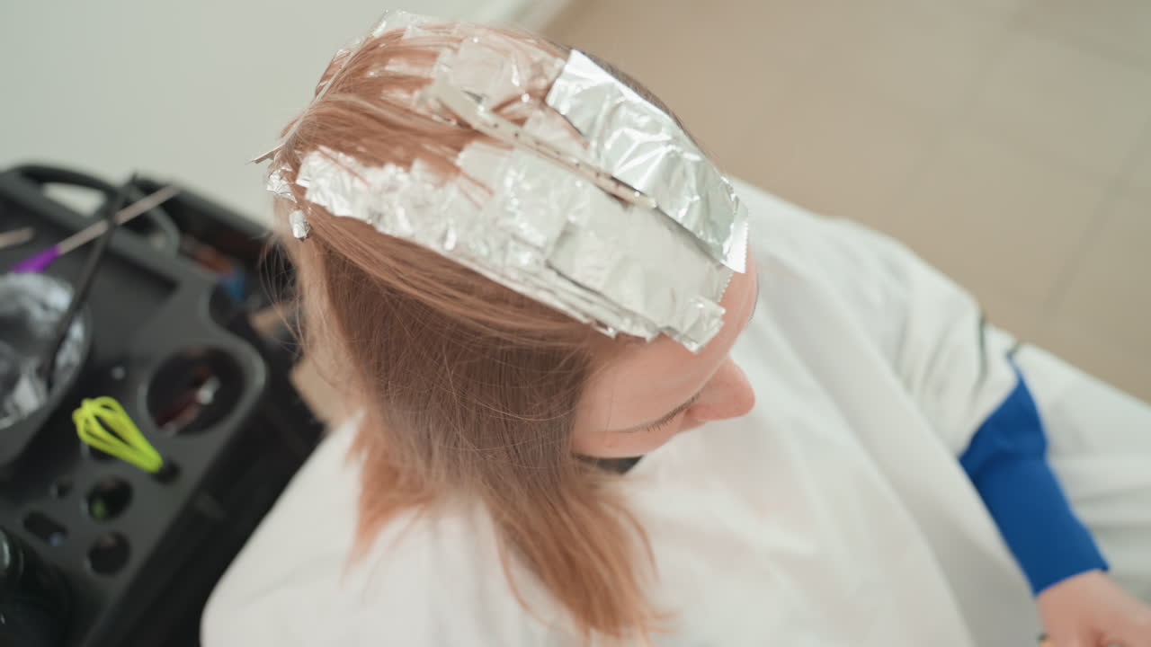Donna caucasica in un salone con stagnola, vista dall'alto di diversi fogli di alluminio che coprono la testa, in attesa durante lo sviluppo del colore, mantellina bianca, vassoio del parrucchiere con strumenti, tenui tonalità bionde che emergono