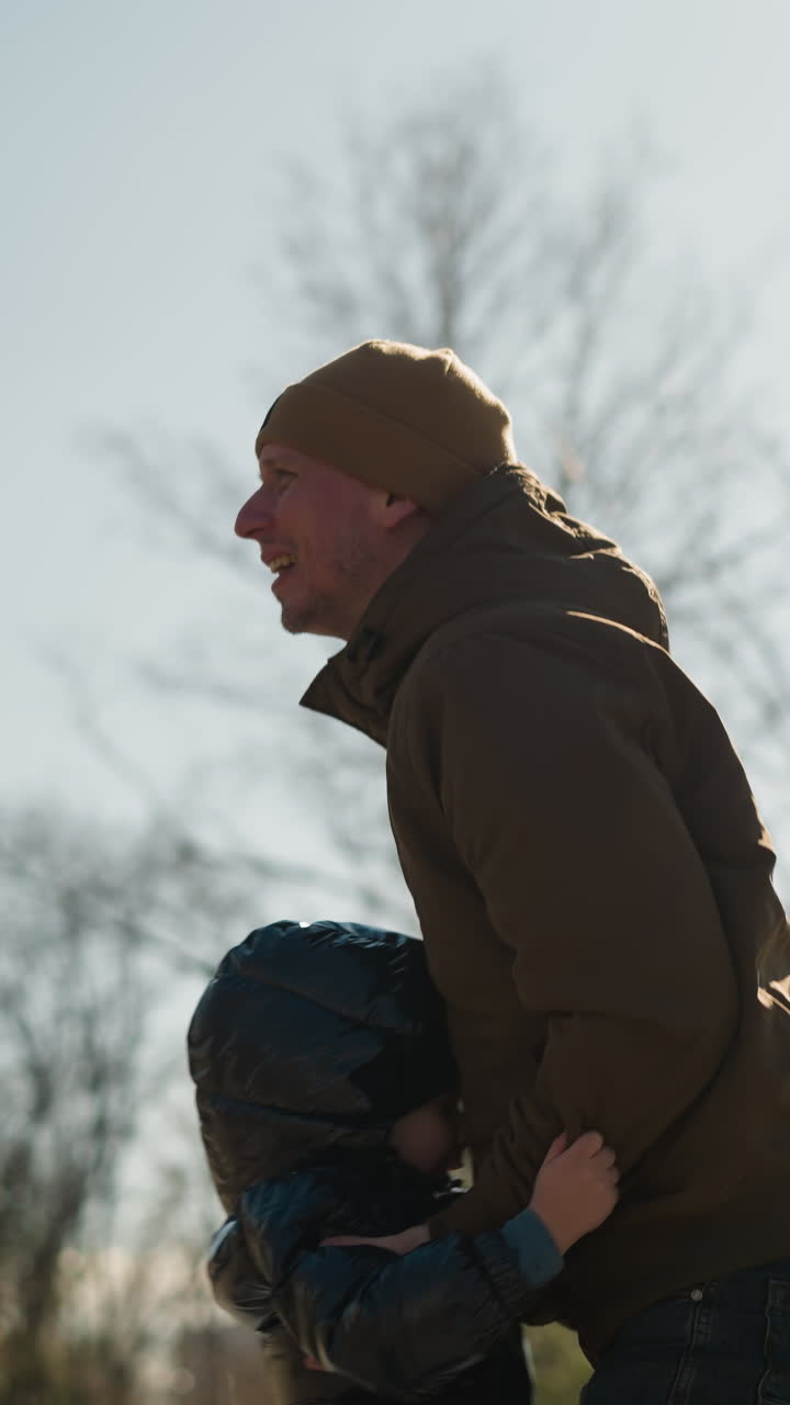Father lifting up his son happily, both are dressed in jeans and with the child wearing a puffy jacket and the man in a beanie, trees and an electric pole in the background