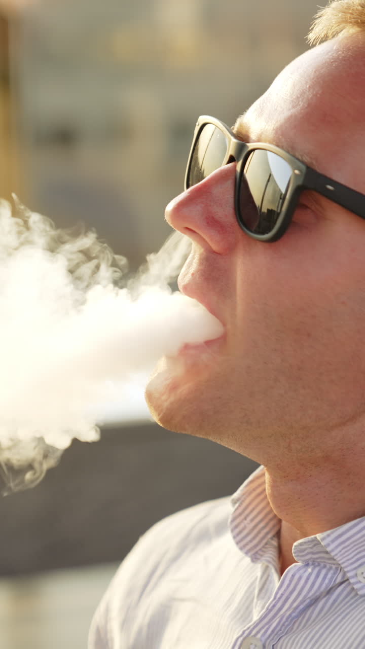 Caucasian young man breathing in smoke from pipe. Male in sunglasses makes a lot of thick white smoke from smoking hookah. Close up. Vertical video